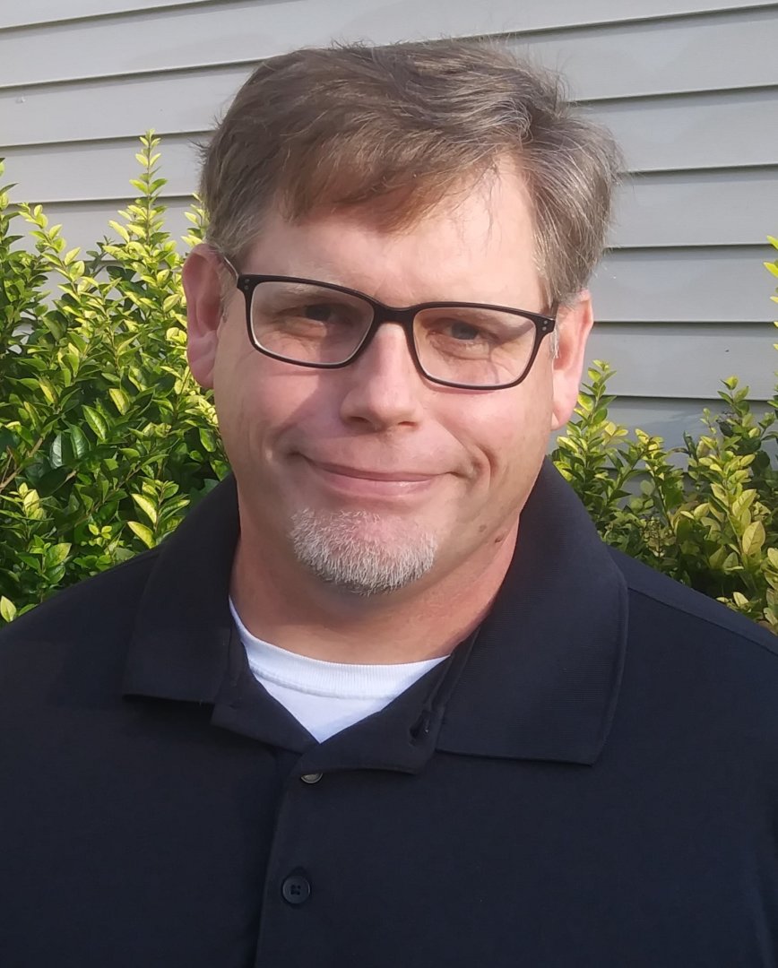 A man with short brown hair, glasses, and a goatee is wearing a black collared shirt and standing outside in front of green bushes and light-colored siding. He is smiling slightly, radiating a caring presence.