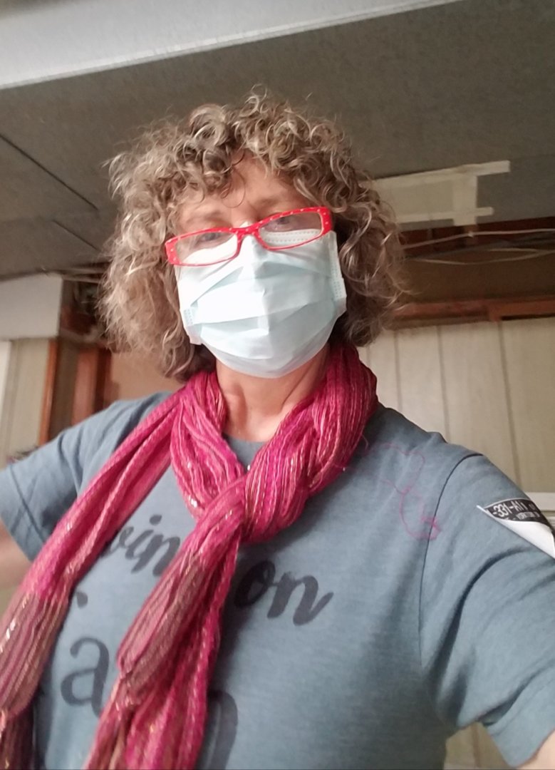 A person with curly hair, red glasses, a face mask, a blue shirt, and a red scarf stands indoors with wood-paneled walls and a ceiling above—reflecting a caring spirit during this powerful time.
