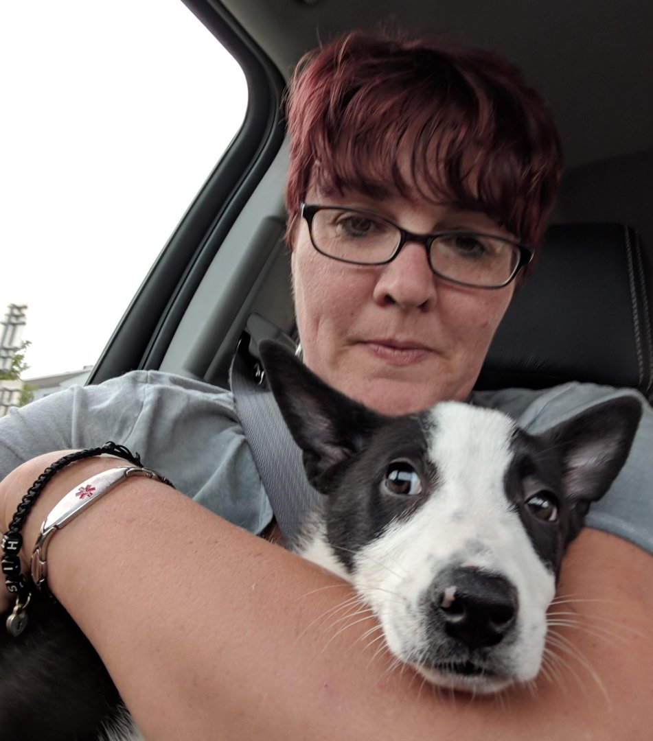 A person with short red hair and glasses, possibly a direct support professional, sits in a car holding a black and white dog close to their chest. Both appear relaxed, and the person is wearing a medical alert bracelet.