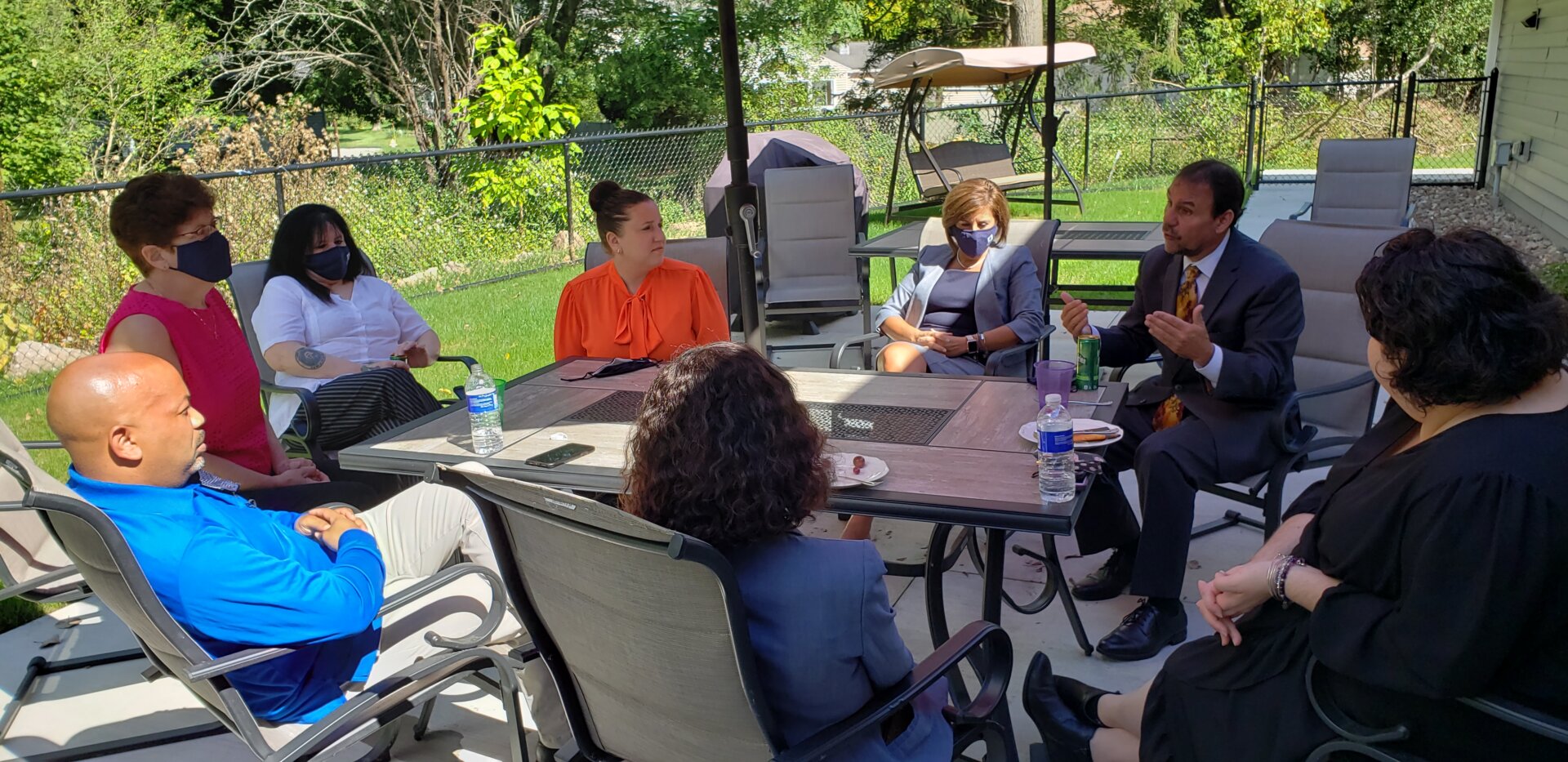 Eight people are seated around a patio table outdoors, engaged in conversation. Some participants are taking notes and one is speaking; one person is wearing a face mask. Trees and a swing set are visible in the background.