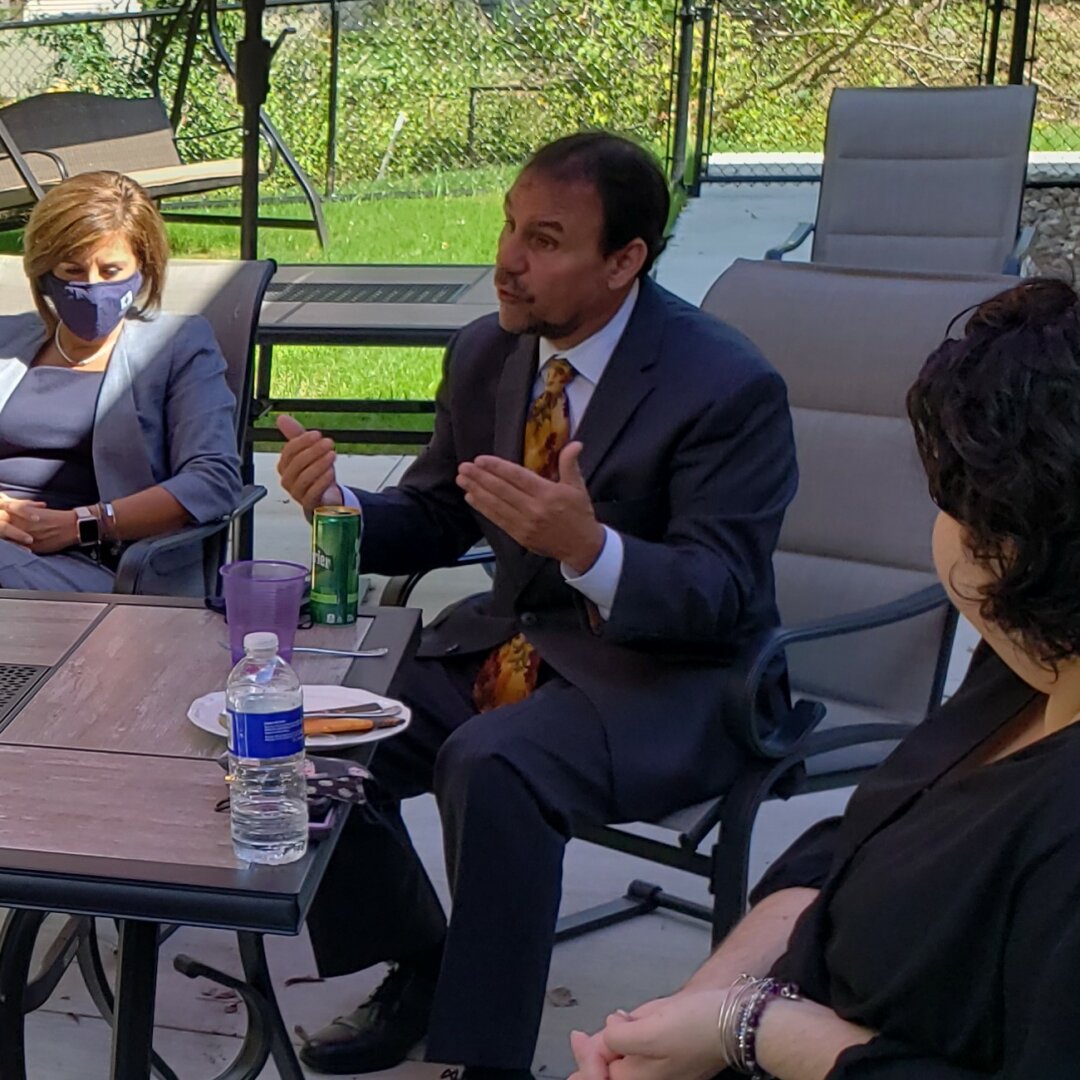 Three people sit outdoors around a table. One man gestures while speaking, as two women listen. One woman wears a face mask. There are drinks and a plate of food on the table, with a grassy area in the background.