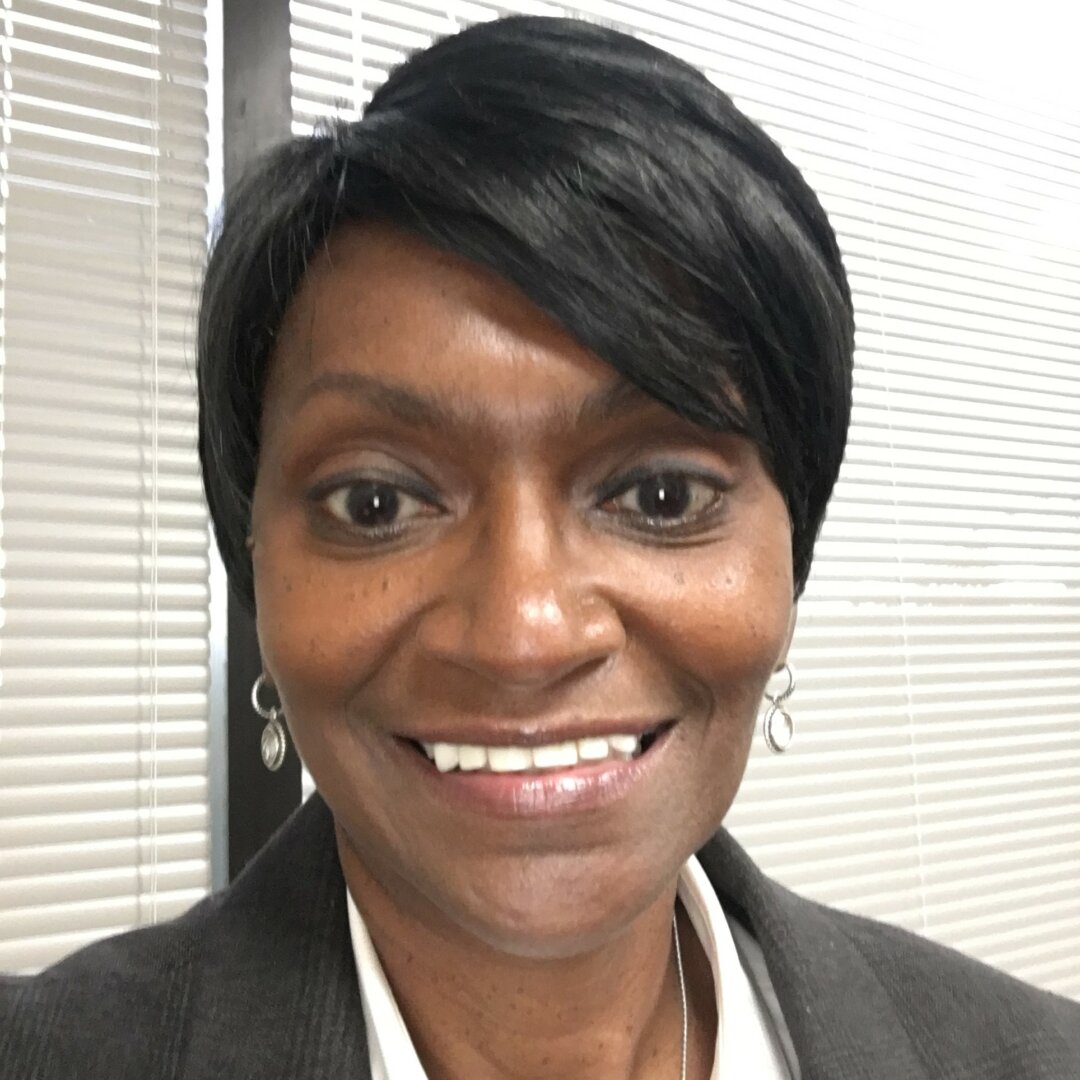 Donna Mostiller, with short black hair, smiles at the camera in a gray blazer, light makeup, and drop earrings. Vertical blinds and a window form the background, setting a professional scene for her Q&A session.
