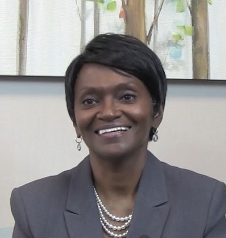 A woman with short black hair, wearing a gray blazer, pearl necklace, and earrings, smiles while sitting indoors. A painting of trees is visible in the background.