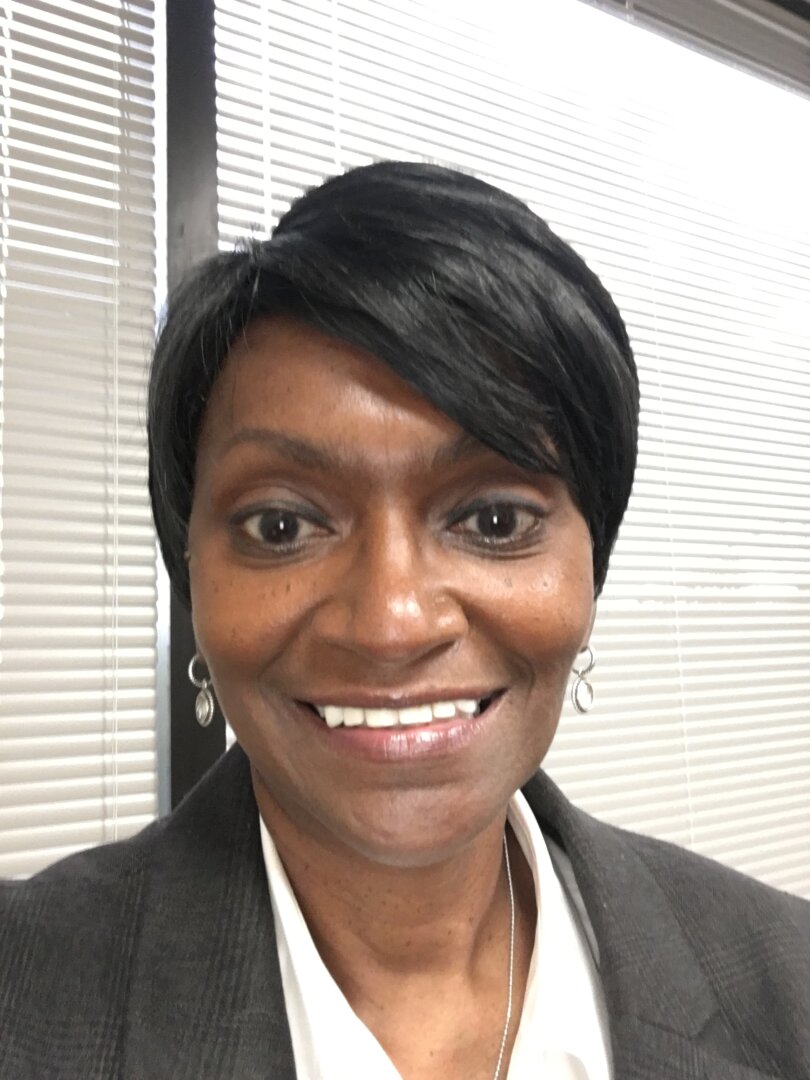 A woman with short black hair, wearing a gray blazer, white shirt, and drop earrings, smiles at the camera in front of window blinds.