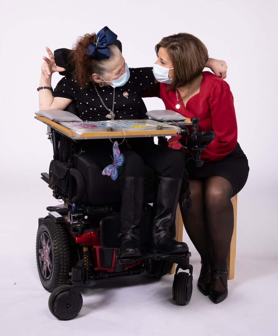 A woman in a red top sits on a stool, leaning toward a woman in a motorized wheelchair. Both wear face masks and appear warmly engaged, reflecting employee commitment and advancing workforce inclusion, with a butterfly decoration on the chair.