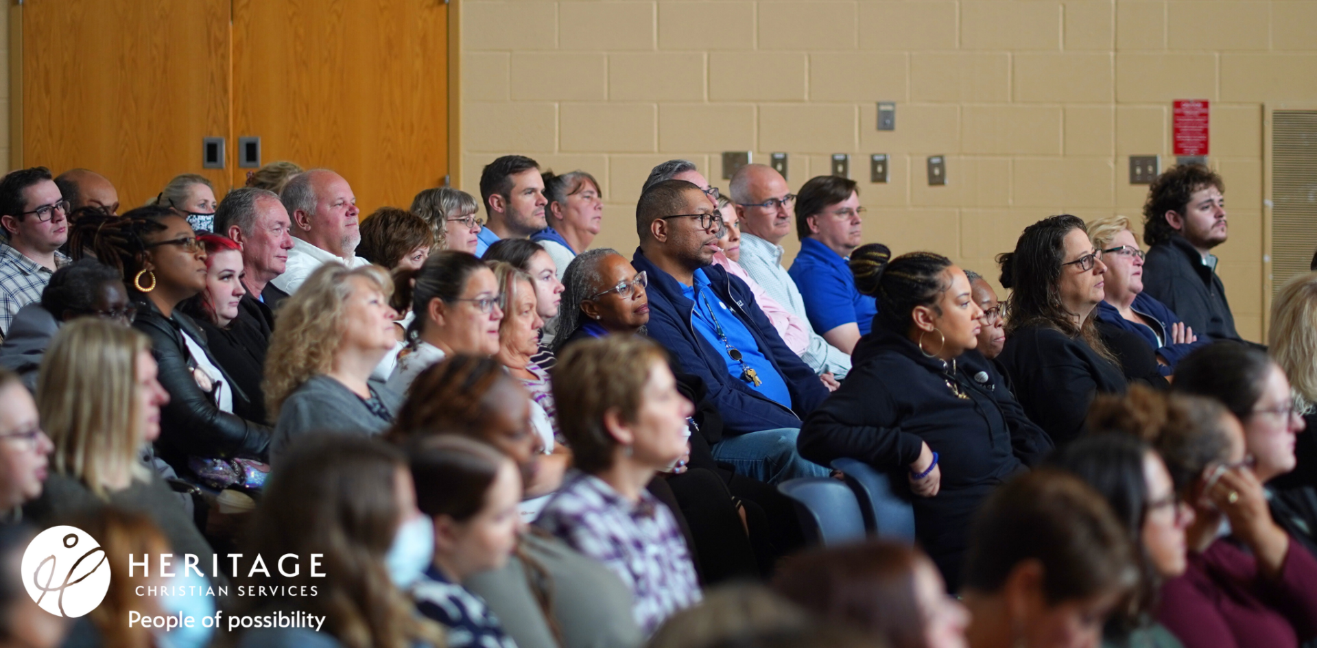 A diverse group of adults attentively listens to a presentation in an auditorium, embodying gratitude and engagement. The Heritage Christian Services logo and the phrase People of possibility appear in the lower left corner.