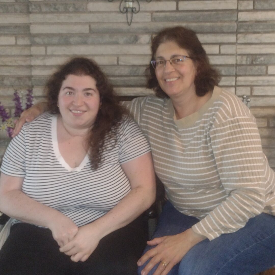 Two women sit close together, smiling at the camera in matching striped shirts. They are seated before a stone wall with a small flower arrangement on the left, sharing a joyful moment as they discuss how to enroll for CDPAP.