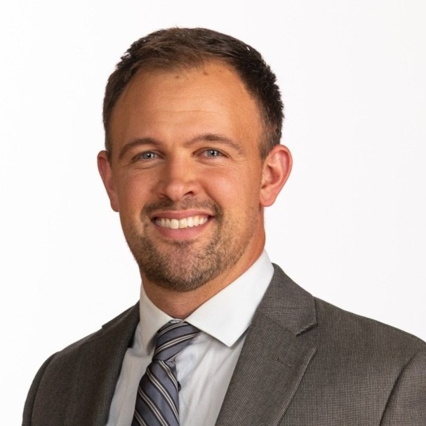 A man with short brown hair and a trimmed beard, wearing a gray suit, white shirt, and striped tie, smiling in front of a plain white background.