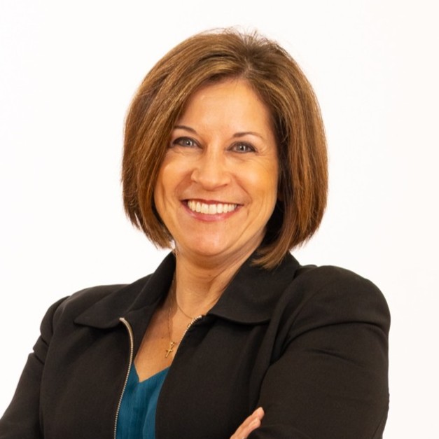 A woman with short brown hair wearing a black blazer and teal top, smiling with arms crossed against a plain white background.
