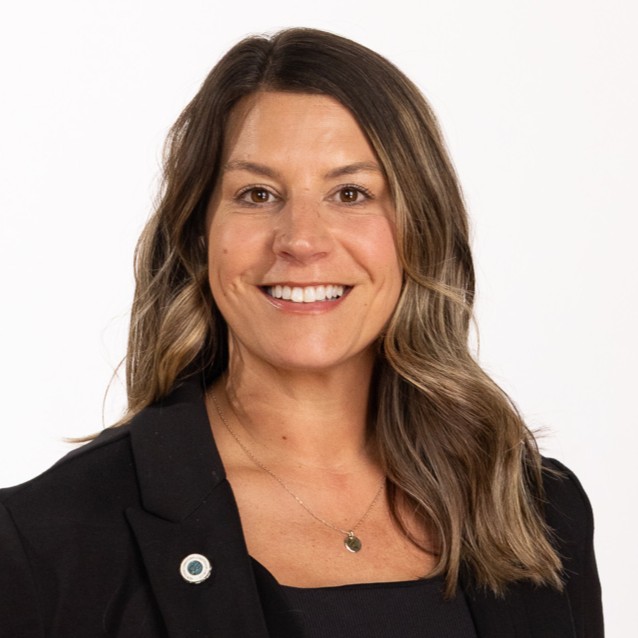 A woman with long brown hair wearing a black blazer, black top, and a necklace, smiling in front of a plain white background.