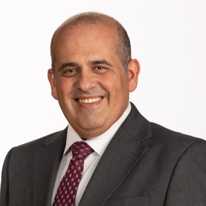 A smiling man with short hair wearing a dark suit, white shirt, and a maroon tie with white patterns, posing in front of a plain white background.