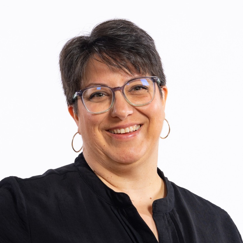 A person with short dark hair, wearing glasses, hoop earrings, and a black shirt, smiles against a plain white background.