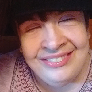 A person with short brown hair, smiling with closed eyes, wearing a patterned light-colored shirt and a dark hat. The background is softly lit and out of focus.