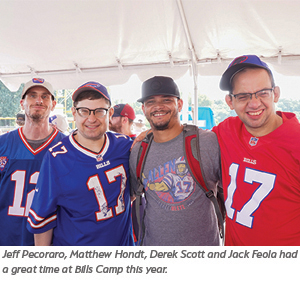 Four men smile and pose together under a white tent at an outdoor event, wearing Buffalo Bills jerseys and caps. A caption below identifies them and mentions they enjoyed Bills Camp this year.