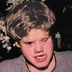 A person with light-colored hair and a patterned shirt is making an expressive face, with one eye closed and lips parted. The background is blurred with white flowers visible.