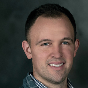 A man with short dark hair, light eyes, and light skin is smiling slightly. He is wearing a checked shirt and a patterned jacket, with a dark, blurred background behind him.