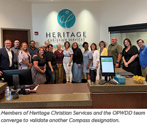 A group of people stand together smiling in the Heritage Christian Services office lobby. The Heritage logo is on the wall behind them. A computer and desk are visible in the foreground.