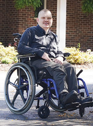 A person with short hair, wearing a dark button-up shirt and pants, sitting and smiling in a blue wheelchair outdoors in front of a brick building and shrubbery.