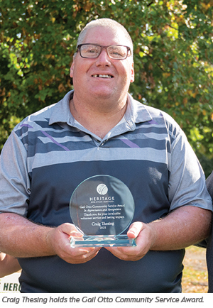 A man wearing glasses and a patterned polo shirt smiles while holding a glass award plaque outdoors. The plaque reads Gall Otto Community Service Award with his name, Craig Thesing, engraved on it.