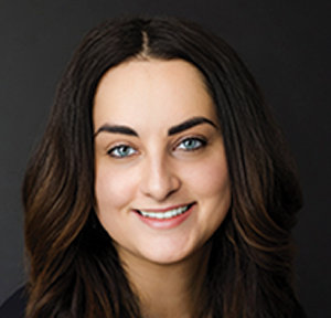A woman with long, wavy brown hair and blue eyes smiles at the camera. She is wearing a dark top and is posed in front of a plain, dark background.