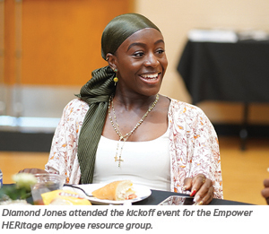A smiling woman wearing a green headscarf sits at a table with food, attending the Empower HERitage employee resource group kickoff event.