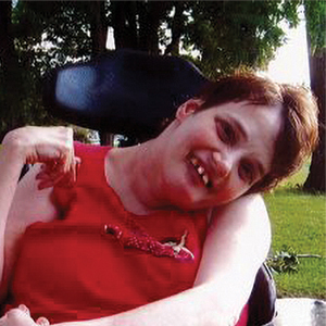 A woman with short brown hair wearing a sleeveless red top sits in a wheelchair outdoors, smiling at the camera. Green grass and trees are visible in the background.