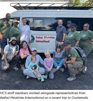 A group of smiling adults and children pose together in front of a van with a Bethel Ministries International sign. Some are kneeling, and others are standing, outdoors on a sunny day.