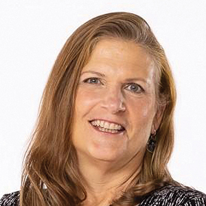 A woman with long, light brown hair smiles at the camera. She is wearing a dark patterned top, earrings, and is photographed against a plain white background.