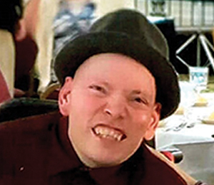 A person wearing a dark hat and a maroon shirt smiles broadly at the camera in a banquet setting, with tables and dishes visible in the background.