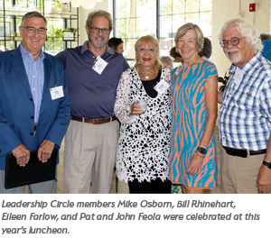 Five adults, three men and two women, stand together indoors, smiling at the camera during a luncheon event. The group is dressed in semi-formal attire and wearing name tags. Bright windows are visible in the background.