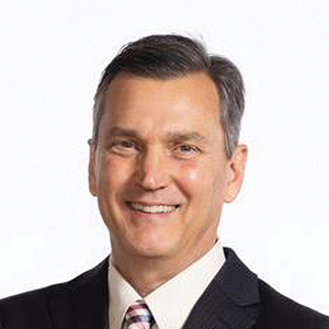 A middle-aged man with short dark hair, wearing a black suit, white shirt, and striped tie, smiling against a plain white background.