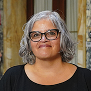 A smiling woman with gray, shoulder-length hair and glasses is wearing a black top. She is standing indoors in front of a marble wall with brown, gold, and white tones.
