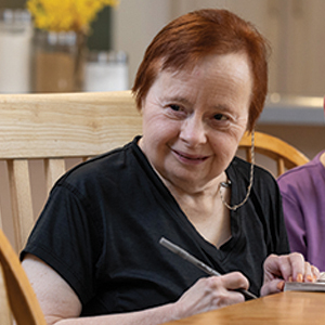 A woman with short red hair sits at a wooden table, smiling and writing in a notebook. She wears a black shirt and is indoors, with a blurred background featuring yellow flowers.