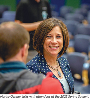 A woman with shoulder-length brown hair smiles while talking to someone at an indoor event, with empty chairs visible in the background.