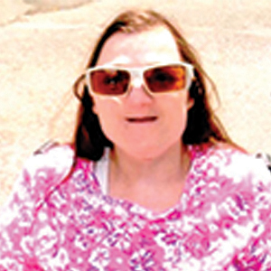 A woman with long brown hair, wearing large sunglasses and a pink patterned top, is smiling outdoors in bright sunlight.