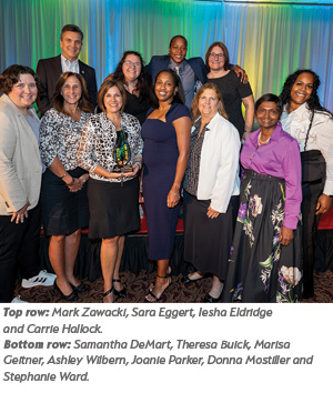 A group of twelve people, dressed in business and semi-formal attire, stand together smiling on a stage with colorful curtains in the background. One woman in the front holds a glass award.