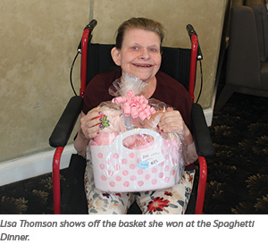 A smiling woman sitting in a wheelchair holds a pink and white gift basket wrapped in cellophane, with a bow on top. She is indoors, and the carpet has a floral pattern.