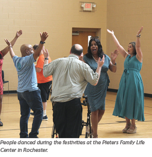 A group of people dance together with raised arms in a gymnasium, celebrating at the Pieters Family Life Center in Rochester. One person uses a walker, while others smile and join in the festivities.