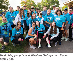 A large group of smiling people wearing matching blue Team Addie shirts pose together outdoors at a fundraising event. Some participants are crouching in front while others stand behind. Trees and a path are visible in the background.