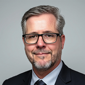 A middle-aged man with gray hair, glasses, and a short beard is wearing a dark suit, light blue shirt, and dark tie, posing against a plain light gray background and smiling slightly.