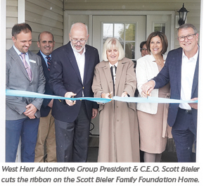 A group of six people, including a woman in a tan coat, stand in front of a house as they cut a blue ribbon at a ceremony. They are smiling, holding large scissors.