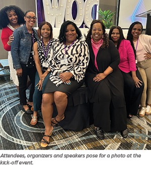 Eight women, both seated and standing, smile and pose together indoors at an event. A large WOC sign is visible behind them, and they are dressed in business casual attire.