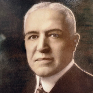 A formal portrait of an older man with short white hair, dressed in a dark suit, white shirt, and patterned tie, looking directly at the camera with a neutral expression. The background is dark and blurred.