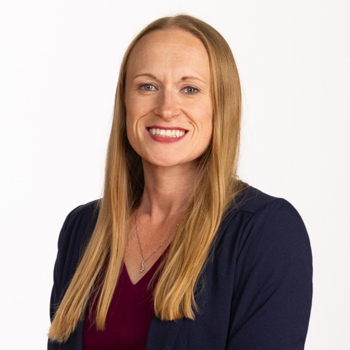 A woman with straight, long blonde hair, wearing a dark blazer over a burgundy top, smiles at the camera against a plain white background.