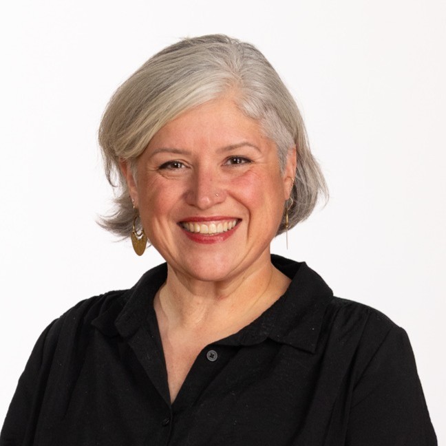 A smiling woman with short gray hair wearing a black shirt and gold earrings poses against a plain white background.
