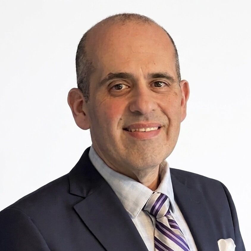 A middle-aged man with short hair and a receding hairline, wearing a navy suit, light-colored shirt, striped tie, and a pocket square, smiling against a plain white background.