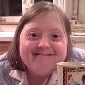 A person with Down syndrome smiles warmly at the camera while sitting indoors, holding a coffee mug decorated with a rooster design. The background includes kitchen counters and dishes.