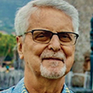 An older man with short gray hair, a trimmed white beard, and glasses smiles outdoors. He is wearing a blue and yellow patterned shirt. The background is blurry, showing greenery and a stone structure.
