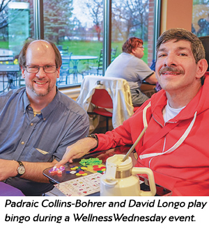 Two men sit at a table with bingo cards and chips, smiling at the camera inside a cafe. Other people are in the background. Text below names them as Padraic Collins-Bohrer and David Longo at a Wellness Wednesday bingo event.