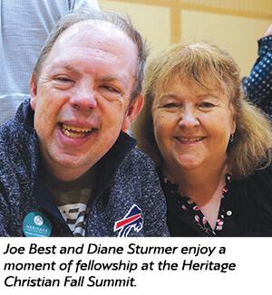 A smiling man and woman sit close together, enjoying each others company at an indoor event. The man wears a Buffalo Bills jacket and a Heritage summit button; the woman has shoulder-length hair and a floral top.